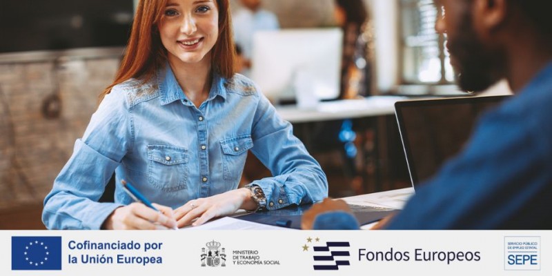 chica peliroja sonriendo sentada de frente a una mesa en un aula con ordenadores y otras personas